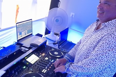 Un DJ souriant mixe de la musique sur une table de mixage avec des ordinateurs dans un environnement lumineux.