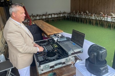 Un DJ mixe de la musique sur une console dans un espace extérieur avec des tables décorées en arrière-plan.