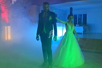 Un couple en tenue de mariage danse dans une salle éclairée par des lumières colorées et entourée de fumée.