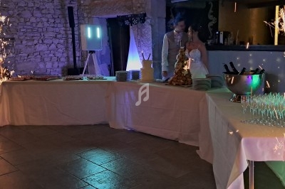 Table de buffet de mariage avec desserts, champagne, et décorations lumineuses, un couple en arrière-plan.
