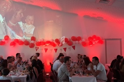 Salle décorée de ballons rouges et blancs, avec des invités assis à des tables et une projection photo sur le mur.