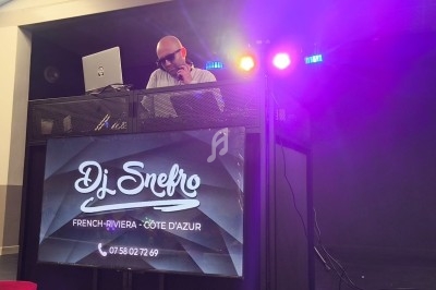 Un DJ mixe sur une scène éclairée par des projecteurs colorés, avec un écran affichant son nom et ses coordonnées.