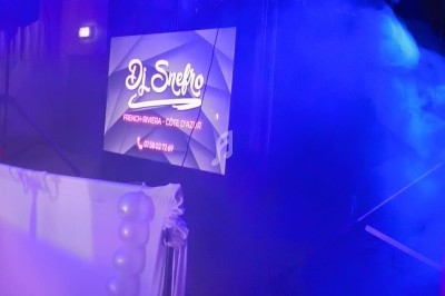 Un DJ mixe sur une scène éclairée de lumières bleues, avec un écran affichant son nom et ses coordonnées.