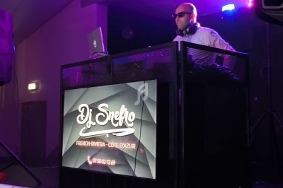 Un DJ mixant sur une scène éclairée par des lumières colorées, derrière une table avec un écran affichant son nom.