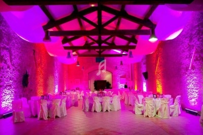Salle de réception avec murs en pierre, éclairage coloré et tables rondes dressées avec nappes blanches.