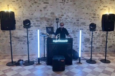 Un DJ mixe derrière une table équipée de matériel audio et lumineux dans une salle en pierre avec un sol carrelé.