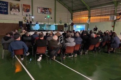 Des personnes assistent à une réunion ou conférence dans un gymnase, avec une table de présentation et un écran projeté.