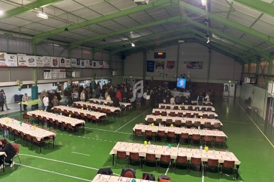 Salle polyvalente avec tables dressées pour un événement, des personnes rassemblées près d'un écran au fond.