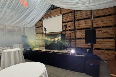 Scène décorée avec rideaux blancs, projecteurs et guirlande colorée, entourée de tables et chaises dans un espace industriel.