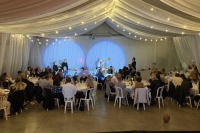 Salle de réception décorée avec des guirlandes lumineuses, des invités assis à des tables rondes et une scène en arrière…