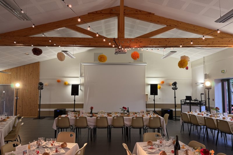 Salle aménagée pour réception avec tables dressées, poutres apparentes, projecteurs et décoration florale suspendue.