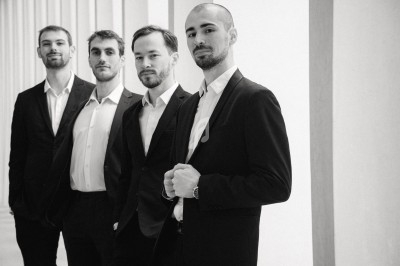 Quatre hommes en costume posant debout dans un espace lumineux avec des colonnes en arrière-plan.