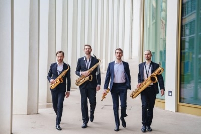 Quatre hommes en costume marchent côte à côte en tenant des saxophones devant un bâtiment moderne.