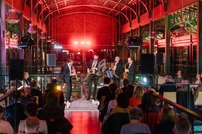 Un groupe de musiciens joue devant un public assis dans un espace intérieur illuminé par des lumières rouges.