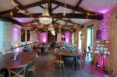 Salle de réception avec poutres apparentes, tables en bois décorées de fleurs et éclairages roses et chaleureux.