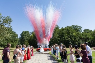 Un couple en tenue de mariage devant une voiture rouge, entouré d'invités, avec des fumées colorées rouge et blanche.