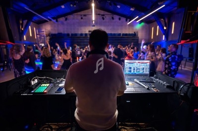 Un DJ mixe de la musique sur une console dans une salle animée où des personnes dansent sous des lumières colorées.