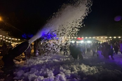 Un canon à mousse projette de la mousse sur une foule lors d'un événement nocturne éclairé par des guirlandes lumineuses.