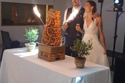 Un couple en tenue de mariage pose devant une pièce montée éclairée par une étincelle, dans une salle décorée simplement.