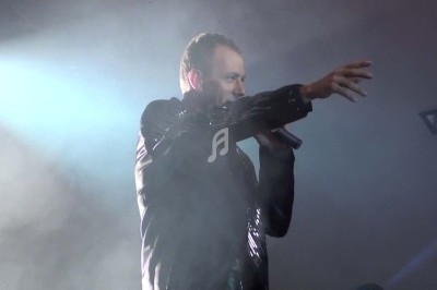 Un chanteur en veste noire tient un micro et pointe du doigt sur une scène éclairée par des projecteurs.