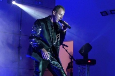 Un chanteur en costume noir brillant se produit sur scène sous un éclairage bleu avec un micro à la main.