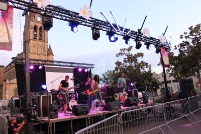 Groupe de musiciens jouant sur une scène extérieure éclairée, avec une église en arrière-plan au crépuscule.