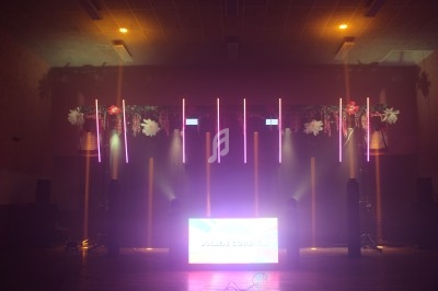 Installation lumineuse avec tubes LED colorés, fleurs suspendues et écran lumineux dans une salle sombre.