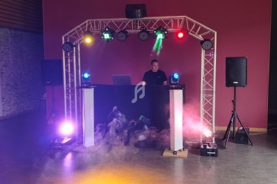 Un DJ mixe derrière une table éclairée par des projecteurs colorés, entouré d'équipements audio et de fumée.