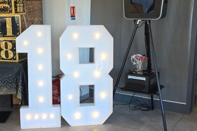 Chiffres lumineux ’18’ décoratifs à côté d'une borne photo dans une salle avec des ballons et une décoration festive.