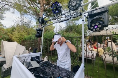 Un DJ mixant en plein air, entouré d'équipements audio et d'éclairages, avec des invités en arrière-plan.