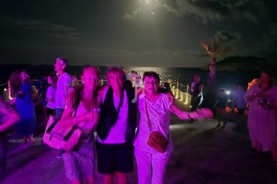 Des personnes dansent et discutent en soirée en plein air sous une lumière violette, avec la mer et la lune en arrière-plan.