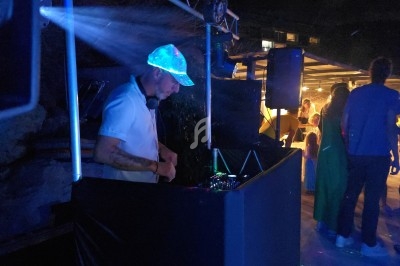 Un DJ mixe de la musique dans un espace extérieur sombre, éclairé par des lumières colorées, avec des personnes en arrière…