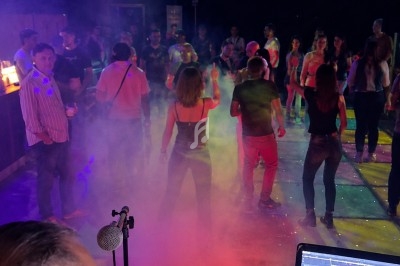 Personnes dansant sur une piste éclairée de lumières colorées, avec un DJ en arrière-plan devant son équipement.