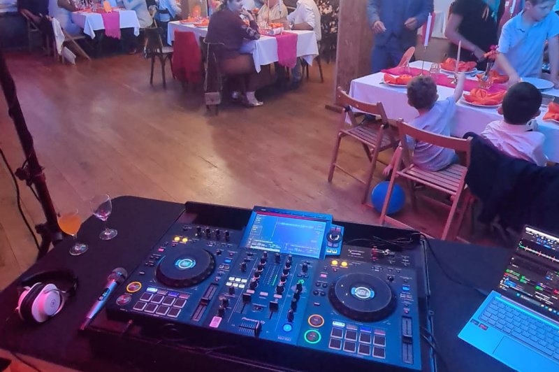 Salle intérieure avec tables dressées, convives attablés, éclairage ambiant et lanterne, console DJ au premier plan.
