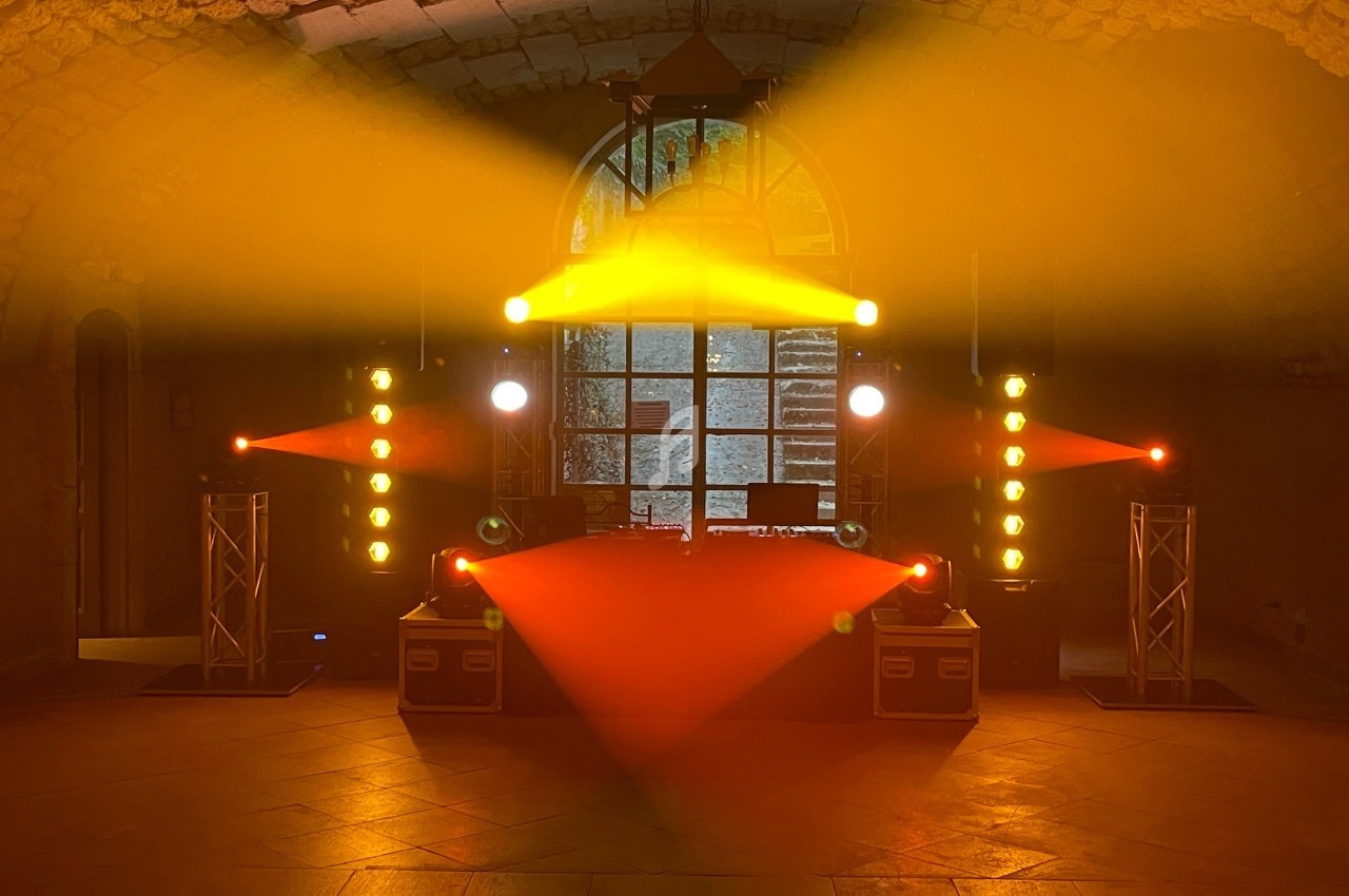 Installation lumineuse avec projecteurs rouges et jaunes dans une salle voûtée en pierre, devant une grande fenêtre.