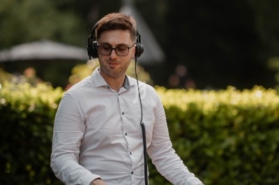 Un DJ en chemise blanche mixe des vinyles en extérieur, entouré de verdure et d'un panneau d'invitation musicale.