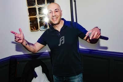 Homme souriant tenant un micro, pointant des doigts, devant un mur clair avec un miroir et un flash lumineux.