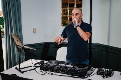 Un DJ en train de mixer et de parler au micro, debout derrière une table équipée de matériel audio.