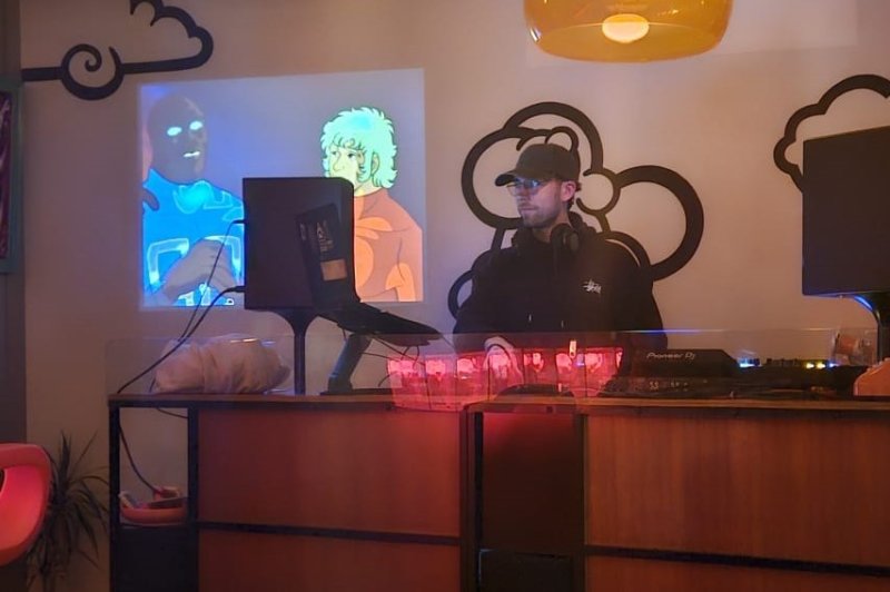 DJ en cabine derrière un pupitre bois, équipement audio visible, écrans d'ambiance colorés, luminaires suspendus dorés, décoration nuages stylisés.