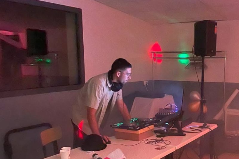 DJ en stand en intérieur face à sa console, éclairage rose et vert, enceintes et équipement autour.